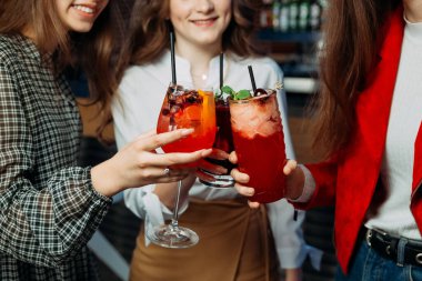 Üç arkadaş bardaklarını canlı kokteyllerle doldurarak arkadaşça kadeh kaldırır ve günlük kıyafetlerini giyerler. Atmosfer canlı ve eğlenceli..