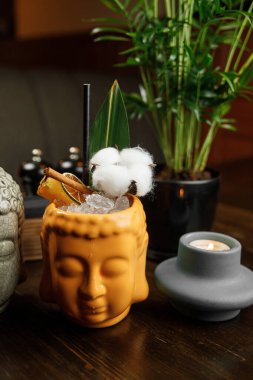 Yaratıcı bir içecek, buzla dolu, pamuk ve bitkiyle kaplı bir Buda kafasında sunulur. Dekoratif unsurlar arasında bir restoran masasında mumlar ve yeşil bitkiler vardır..