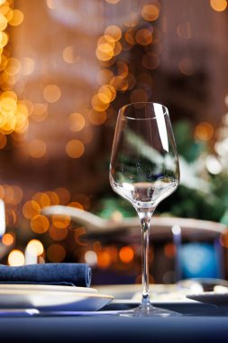 Yemek için setin üzerinde bir şarap kadehi duruyor. Arka planda sıcak, pırıl pırıl ışıklar yer alır ve neşeli bir dokunuş eklenir. Sahne samimi bir yemek deneyimi öneriyor, toplanmak için mükemmel..