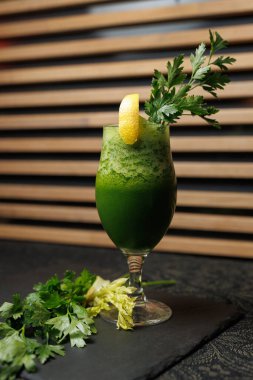 Yakınlarda maydanoz ve kereviz bulunan siyah taştan bir yüzeye serinletici yeşil bir içecek konur. Bu içecek bir limon dilimi içeriyor ve öğle yemeği sırasında trend bir kafede servis ediliyor..
