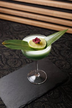 Yeşil bir kokteyl, üzerinde limon dilimi ve dekoratif yapraklar olan uzun bir bardakta servis edilir. İçkiler siyah bir tabağa konur ve şık bir arka plana yerleştirilir..