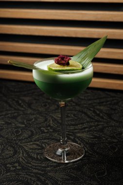Uzun bir bardakta renkli bir kokteyl servis edilir. İçecekte yeşil katmanlar, bir dilim limon ve bir yaprak süslemesi yer alıyor. Dekorun arka planında tahta çıta olan koyu renkli bir masası var..