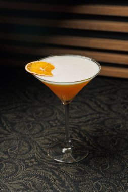 Bir kokteyl, kenarında bir dilim portakal olan uzun bir bardakta zarif bir şekilde durur. İçkinin üzerinde açık renk ve kremalı köpük var..