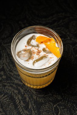 Buzla dolu berrak bir bardak ve portakal dilimli bir içecek karanlık bir yüzeye oturur. İçkiler ferahlatıcı görünüyor, sıcak hava ve sosyal etkinlikler için ideal..