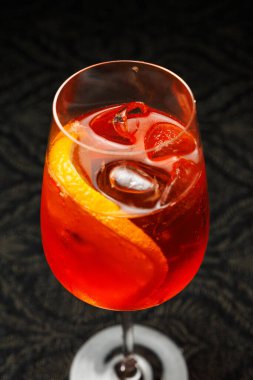 The drink features a mixture of vibrant colors with ice cubes and a fresh orange slice placed on the rim of the glass. It is set against a dark background, perfect for an evening out.