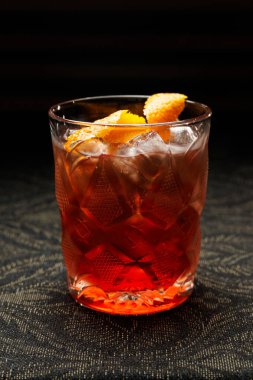 A drink is served in a textured glass filled with ice and garnished with orange peels. The dark background highlights the vibrant color of the beverage.