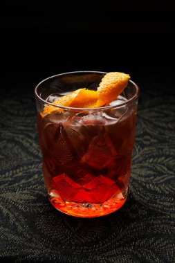 A glass filled with a colorful cocktail rests on a dark patterned table. The drink has ice and is garnished with a twist of orange peel. The setting is cozy and inviting.