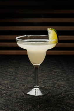 İçki bir bardağın içinde tuz çemberi ve kenarında limon dilimi ile duruyor. Koyu renk ahşap masa içkinin açık rengine zıtlık ekler. Bu sahne davetkar bir restoran atmosferinde geçiyor..