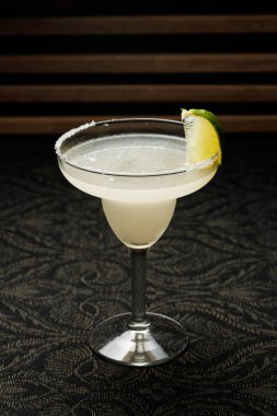 Margarita, tuzlu kenarlı ve yanında limon dilimi olan berrak bir bardakta servis edilir. İçki, gece boyunca bir barda, koyu desenli bir masanın üzerine konur..