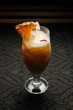 A tall glass showcases a tropical cocktail filled with ice. The drink has a garnish of pineapple and coconut. The background features a textured dark surface, creating a warm atmosphere.