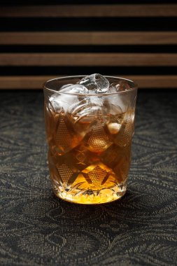 A clear glass filled with a classic drink sits on a dark patterned surface. Ice cubes fill the glass, reflecting light as the drink looks inviting and refreshing.