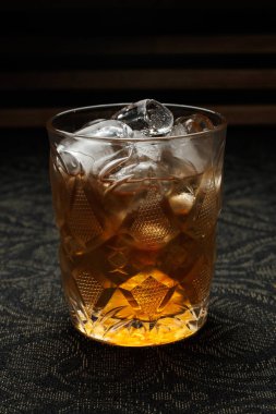 A drink filled with ice sits in a decorative glass on a dark surface, creating a lively setting for relaxation. This drink offers a perfect choice for evening social events.