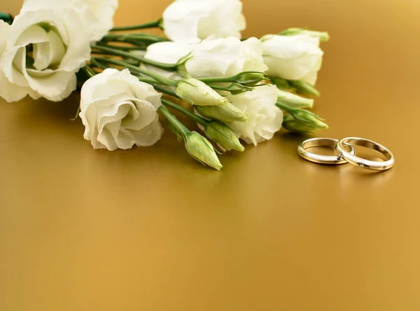金の背景のストック画像上の白い花と2つの黄金の結婚指輪 白い花のイメージの花束で婚約指輪 テキストのコピースペースとロマンチックな黄金の結婚式の背景 ウェディングリングフレーム — ストック写真