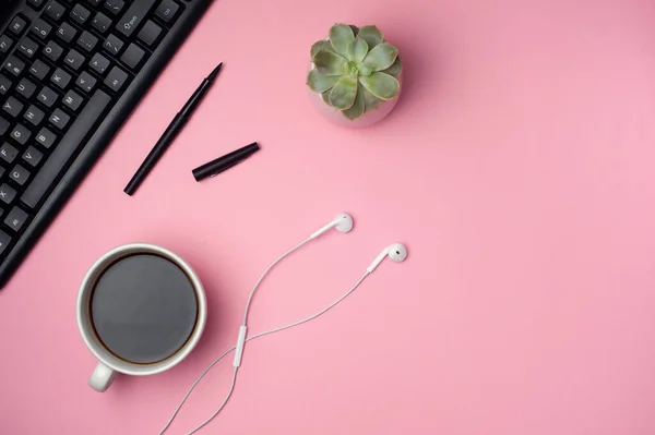 Minimal office desk. Flat lay - keyboard, white earphones, cup of coffee on pink background. Top view. Workplace concept.