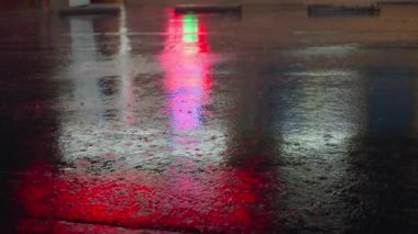 Ağır çekim yağmur damlaları geceleyin asfaltta birikintiye düşer. Büyük modern şehrin ışıkları su yüzeyine yansıyor. Yağmur mevsiminde yağmur yağarken yakın çekim. Yağmur damlaları yağıyor.