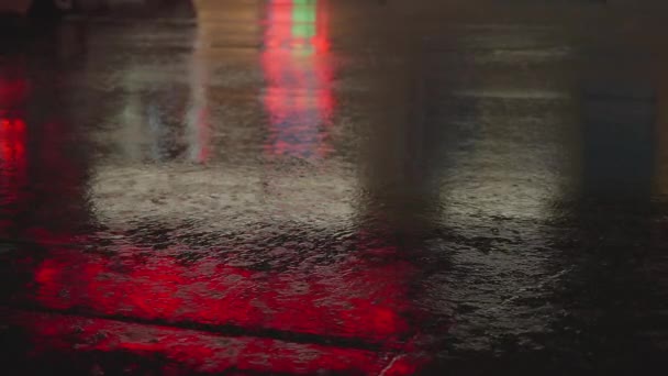 Regendruppels lopen door het glas van de auto tegen de achtergrond van de felle lichten van de nachtstad. Een regenachtige nacht. Auto 's passeren het kruispunt. Helder gekleurde verkeerslichten. — Stockvideo