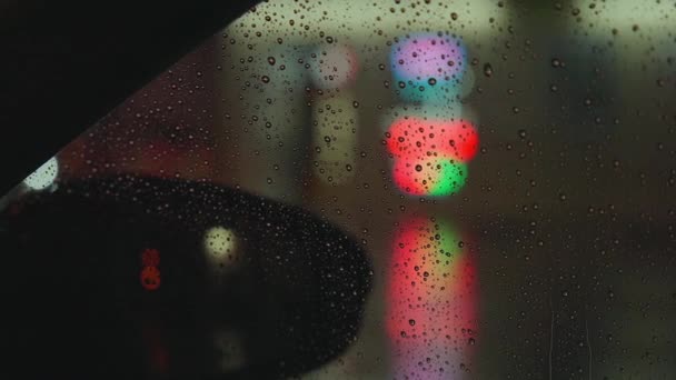 Gotas de lluvia corren por el cristal del coche contra el telón de fondo de las luces brillantes de la ciudad nocturna. Noche lluviosa. Los coches pasan la intersección. Semáforos de colores brillantes. — Vídeos de Stock