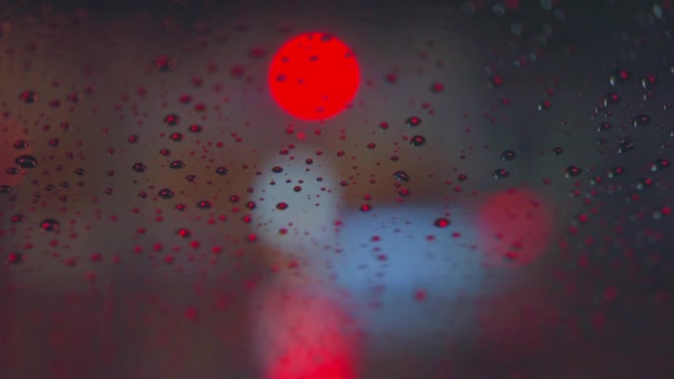 Az esőcseppek végigfutnak az autó üvegén az éjszakai város fényes fényeinek hátterében. Esős éjszaka. Autók haladnak át a kereszteződésen. Világos színű közlekedési lámpák. — Stock videók