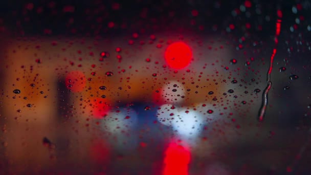 Gotas de lluvia corren por el cristal del coche contra el telón de fondo de las luces brillantes de la ciudad nocturna. Noche lluviosa. Los coches pasan la intersección. Semáforos de colores brillantes. — Vídeo de stock