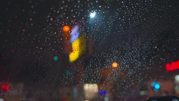 Krople deszczu spływają po szybie samochodu na tle jasnych świateł nocnego miasta. Deszczowa noc. Samochody przejeżdżają obok skrzyżowania. Jasne kolorowe światła drogowe. — Wideo stockowe