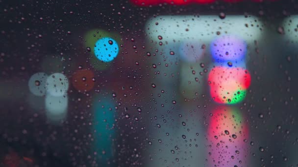 Gotas de lluvia corren por el cristal del coche contra el telón de fondo de las luces brillantes de la ciudad nocturna. Noche lluviosa. Los coches pasan la intersección. Semáforos de colores brillantes. — Vídeos de Stock