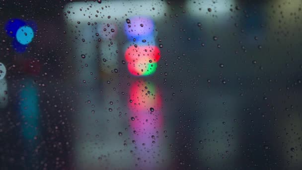 Gotas de chuva correm pelo vidro do carro contra o pano de fundo das luzes brilhantes da cidade noturna. Noite chuvosa. Os carros passam pelo cruzamento. semáforos coloridos brilhantes. — Vídeo de Stock