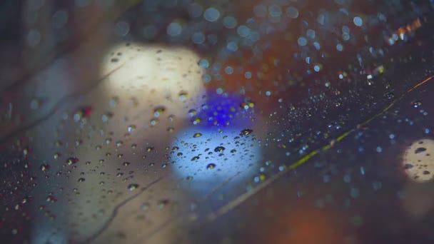 Regndroppar rinner ner i glaset av bilen mot bakgrund av de ljusa lamporna i nattstaden. Regnig kväll. Bilar passerar korsningen. Ljusfärgade trafikljus. — Stockvideo