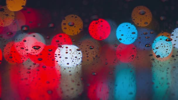 Gotas de lluvia corren por el cristal del coche contra el telón de fondo de las luces brillantes de la ciudad nocturna. Noche lluviosa. Los coches pasan la intersección. Semáforos de colores brillantes. — Vídeos de Stock