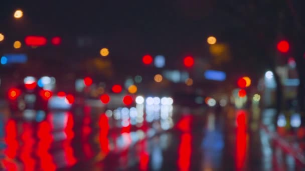 Az esőcseppek végigfutnak az autó üvegén az éjszakai város fényes fényeinek hátterében. Esős éjszaka. Autók haladnak át a kereszteződésen. Világos színű közlekedési lámpák. — Stock videók