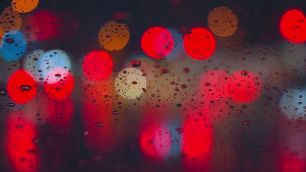 Yağmur damlaları arabanın camından gece şehrinin parlak ışıklarının arka planına doğru akıyor. Yağmurlu bir gece. Arabalar kavşağı geçsin. Parlak renkli trafik ışıkları. — Stok video