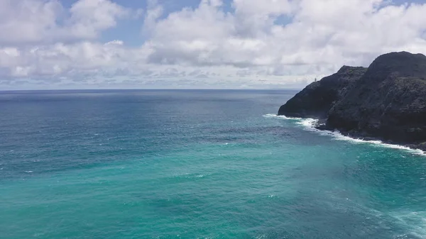 Latający dron nad oceanem. Widok na latarnię morską Makapuu. Fale Oceanu Spokojnego myją skalisty brzeg. Wspaniałe góry hawajskiej wyspy Oahu na tle błękitnego nieba z białymi chmurami. — Zdjęcie stockowe