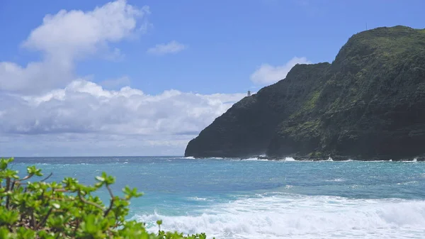 Widok na latarnię morską Makapuu. Fale Oceanu Spokojnego spływają po żółtym piasku tropikalnej plaży. Wspaniałe góry hawajskiej wyspy Oahu na tle błękitnego nieba z białymi chmurami. — Zdjęcie stockowe