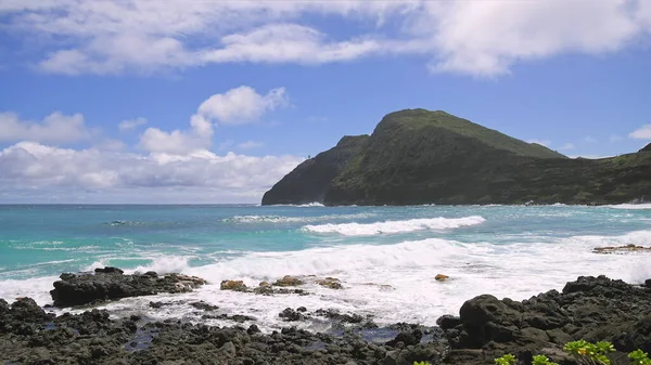 Άποψη του φάρου makapuu. Τα κύματα του Ειρηνικού Ωκεανού πλένονται πάνω από την κίτρινη άμμο της τροπικής παραλίας. Μεγαλοπρεπή βουνά της Χαβάης νησί του Oahu με φόντο το γαλάζιο του ουρανού με λευκά σύννεφα. — Φωτογραφία Αρχείου