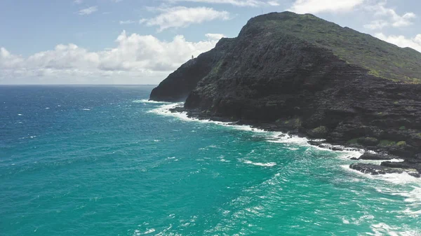 Drone volante sull'oceano. Veduta del faro di Makapuu. Onde dell'Oceano Pacifico lavano la costa rocciosa. Magnifiche montagne dell'isola hawaiana di Oahu sullo sfondo del cielo blu con nuvole bianche. — Foto Stock
