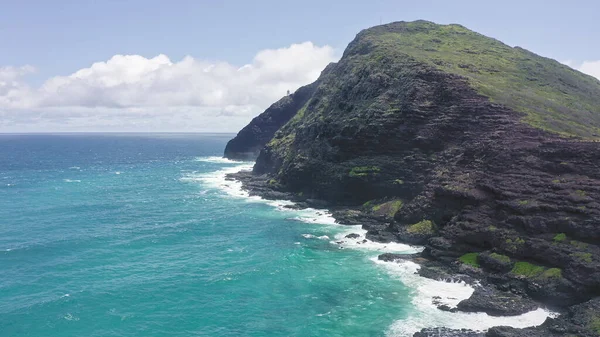 Um drone voador sobre o oceano. Vista do farol makapuu. Ondas de Oceano Pacífico lavam a costa rochosa. Magníficas montanhas da ilha havaiana de Oahu contra o pano de fundo do céu azul com nuvens brancas. — Fotografia de Stock