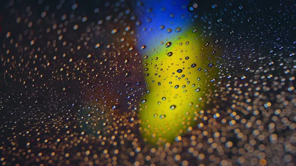 Gotas de chuva correm pelo vidro do carro contra o pano de fundo das luzes brilhantes da cidade noturna. Noite chuvosa. Os carros passam pelo cruzamento. semáforos coloridos brilhantes. — Fotografia de Stock