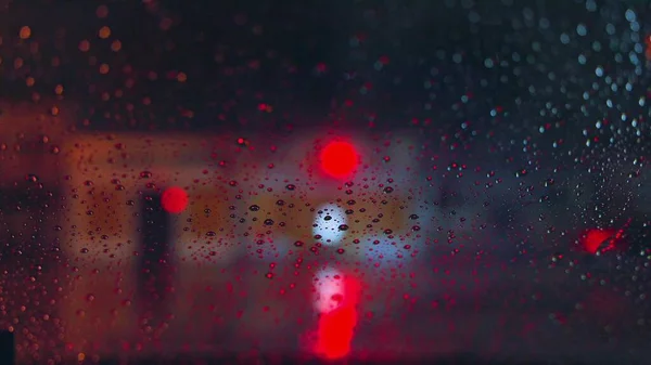 Regendruppels lopen door het glas van de auto tegen de achtergrond van de felle lichten van de nachtstad. Een regenachtige nacht. Auto 's passeren het kruispunt. Helder gekleurde verkeerslichten. — Stockfoto