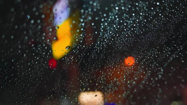 Gotas de chuva correm pelo vidro do carro contra o pano de fundo das luzes brilhantes da cidade noturna. Noite chuvosa. Os carros passam pelo cruzamento. semáforos coloridos brilhantes. — Fotografia de Stock