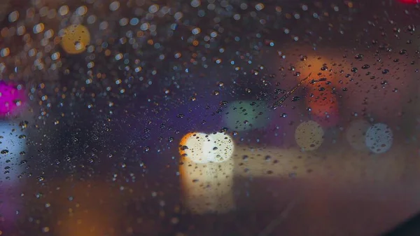 Gotas de chuva correm pelo vidro do carro contra o pano de fundo das luzes brilhantes da cidade noturna. Noite chuvosa. Os carros passam pelo cruzamento. semáforos coloridos brilhantes. — Fotografia de Stock