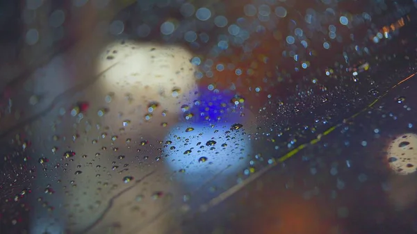 Vor dem Hintergrund der hellen Lichter der nächtlichen Stadt fließen Regentropfen durch die Scheibe des Autos. Verregnete Nacht. Autos passieren die Kreuzung. Knallbunte Ampeln. — Stockfoto