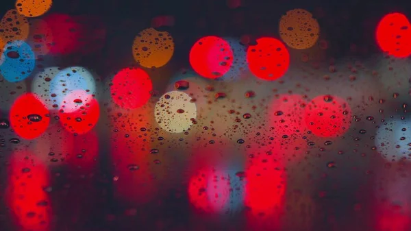 Gotas de chuva correm pelo vidro do carro contra o pano de fundo das luzes brilhantes da cidade noturna. Noite chuvosa. Os carros passam pelo cruzamento. semáforos coloridos brilhantes. — Fotografia de Stock