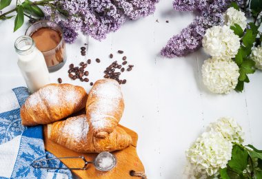 Taze kruvasanlar, bardak kahve, süt, çilek reçeli ve Beyaz ahşap zemin üzerinde çiçekler ile Kahvaltı