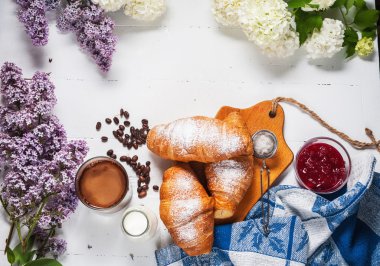 Taze kruvasanlar, bardak kahve, süt, çilek reçeli ve Beyaz ahşap zemin üzerinde çiçekler ile Kahvaltı