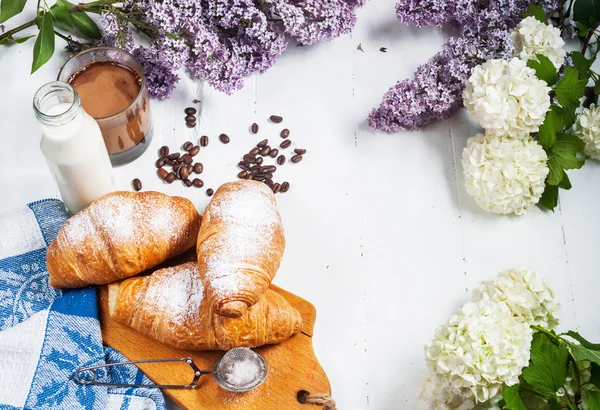 Taze kruvasanlar, bardak kahve, süt, çilek reçeli ve Beyaz ahşap zemin üzerinde çiçekler ile Kahvaltı