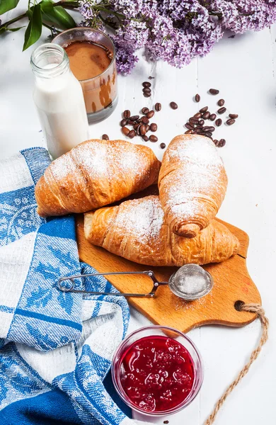 Taze kruvasanlar, bardak kahve, süt, çilek reçeli ve Beyaz ahşap zemin üzerinde çiçekler ile Kahvaltı