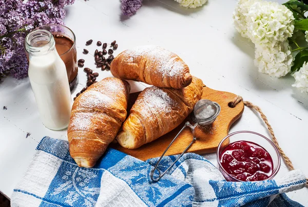 Taze kruvasanlar, bardak kahve, süt, çilek reçeli ve Beyaz ahşap zemin üzerinde çiçekler ile Kahvaltı