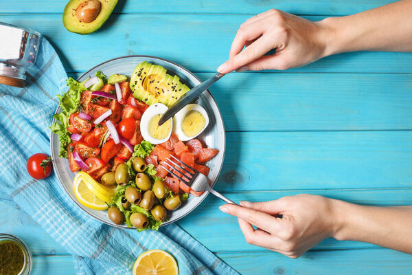 Ketogenic, paleo diet lunch bowl with salted salmon fish, lemon, avocado, olives, boiled egg, tomato, green lettuce salad. Female hands holds fork and knife. top view