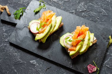 Koyu çavdar ekmeği, avokado, tütsülenmiş somon, salatalık ve turplu siyah taş zemin. Üst görünüm