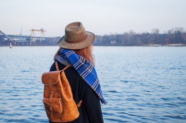 Genç kadın şapkalı ve nehir kıyısındaki ayakta, gündoğumu veya günbatımı ufka, geri görünüm seyir sırt çantası ile