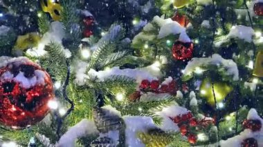 Noel ağacı dışarıda yağan karın altında oyuncaklar ve çelenklerle süslenir. Yeni yıl geçmişi çok renkli. Akşamları yanan ışıklar. 4K Canlandırma Videosu.